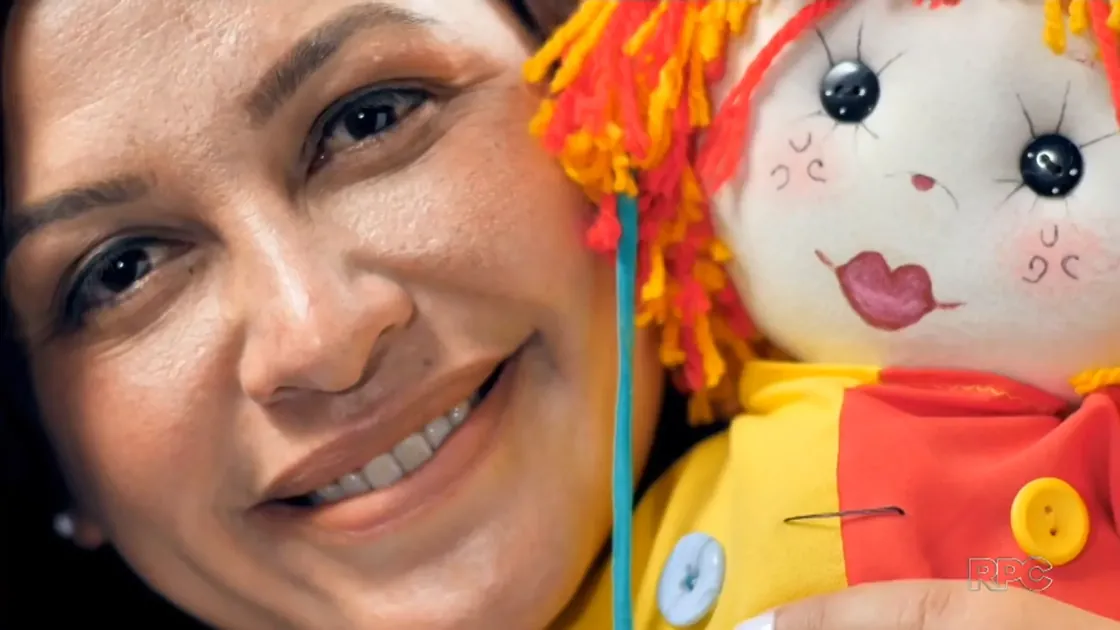 Tia Edi sorrindo ao lado de uma boneca artesanal, em reportagem da RPC.
